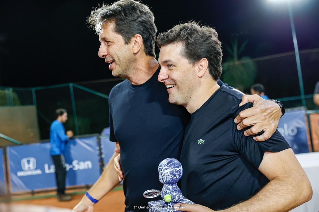 Dois tenistas abraçados na quadra de saibro à noite, um deles segurando o troféu do campeão