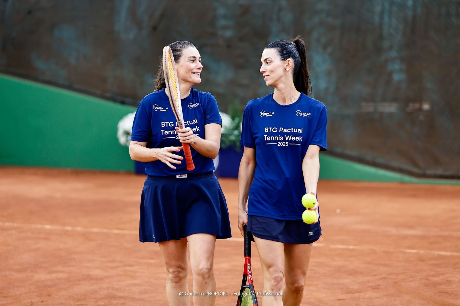 Duas jogadoras com camisa da BTG Pactual Tennis Week 2025 na quadra de saibro